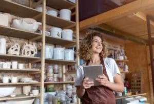 A woman wearing a white t-shirt and a maroon apron smiles and holds a business tablet in a pottery studio with shelves of products in the background.