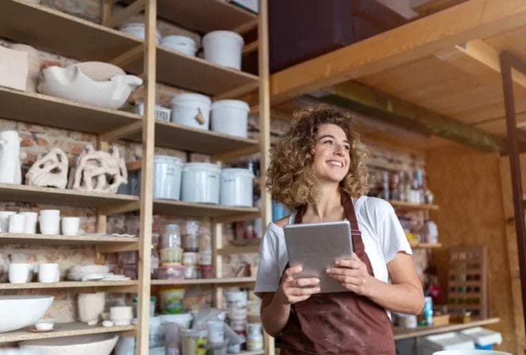 A woman wearing a white t-shirt and a maroon apron smiles and holds a business tablet in a pottery studio with shelves of products in the background.