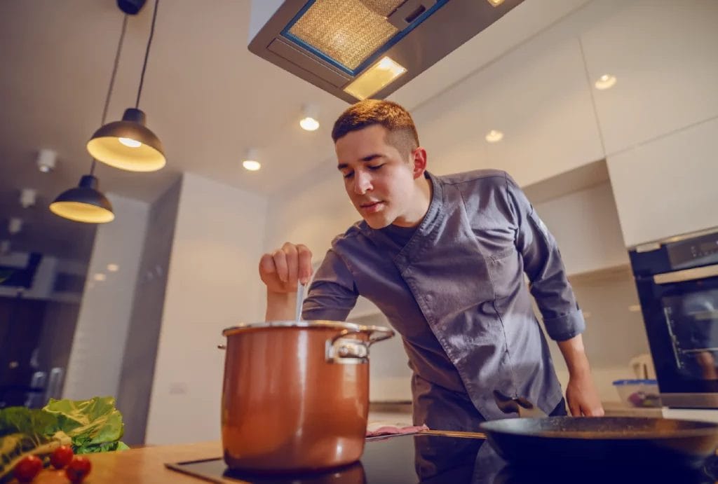 A private chef wearing a gray chef's coat sprinkles an ingredient into a large copper pot on an induction stove in a bright modern kitchen.