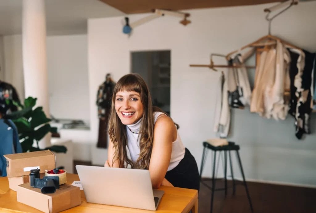 A woman with long brown hair, wearing a sleeveless white top, smiles and rests her elbows on a wooden table in front of an open laptop, shipping tape, and brown shipping boxes.