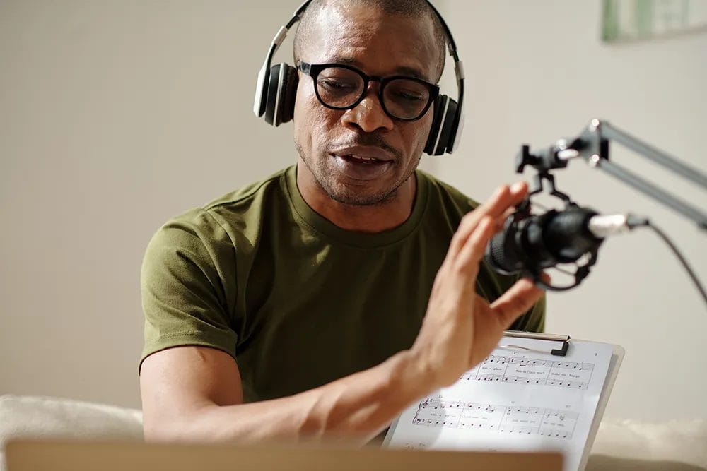 Online music teacher wearing headphones and holding up a clipboard with sheet music speaks into a microphone