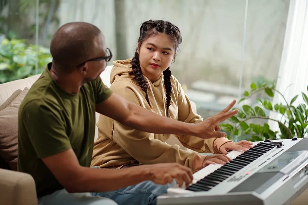 Home music teacher explains a concept to a high school student while playing on a keyboard.