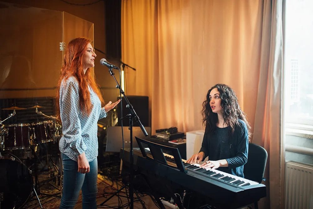 Student sings into a microphone in a vocal lesson while a music teacher plays the piano and gives her notes