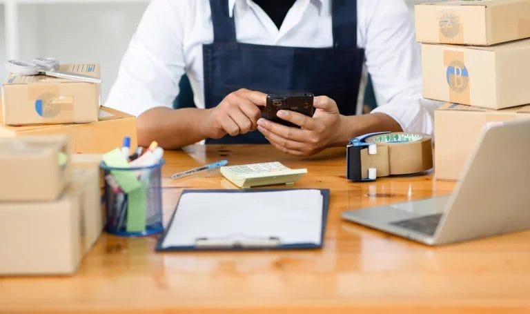 A small business owner using a smart phone at a desk, surrounded by shipping boxes, packing tape, a laptop, and office supplies.