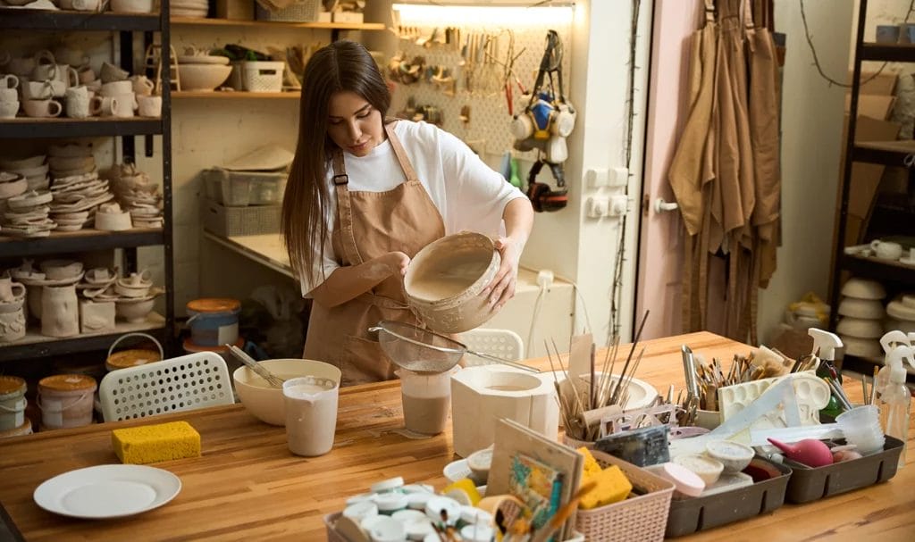 A potter is carefully pouring glazing liquid through a strainer in her studio.