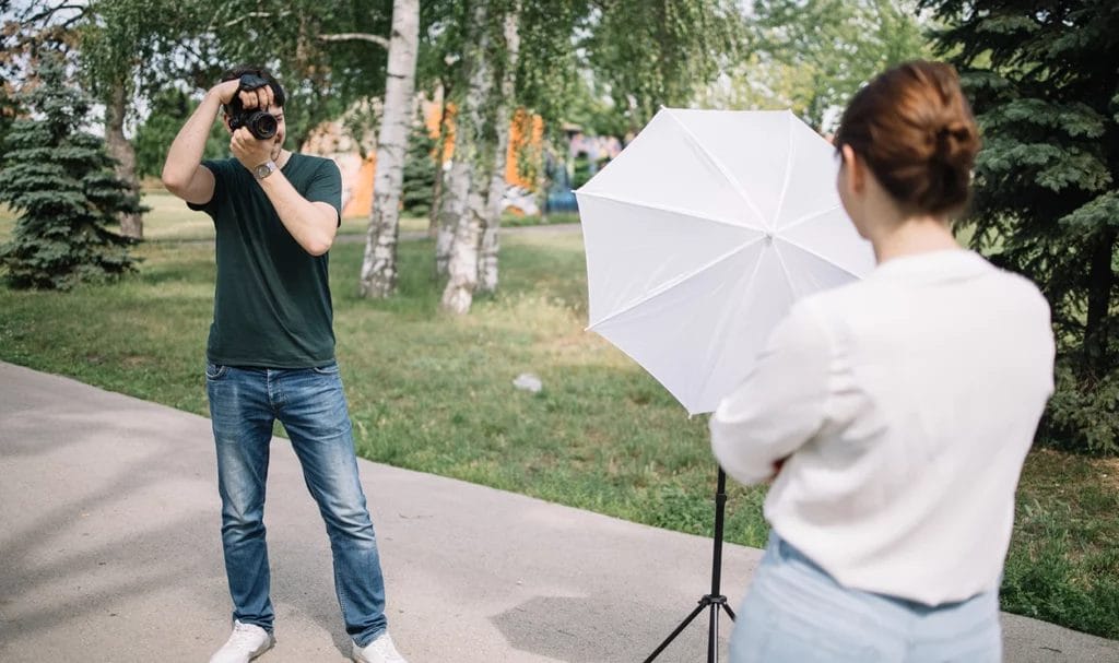 A photographer is taking a photo of their client, who is posed next to a lighting tripod in a public park.