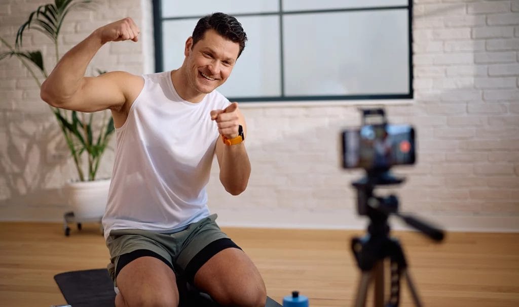 A personal trainer kneeling on a yoga mat next to some weights in an indoor exercise studio, flexing a pose for a livestream as he points to his smart phone on a tripod.
