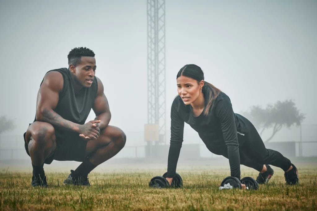 A trainer crouches next to a client performing renegade rows outside with a foggy background.