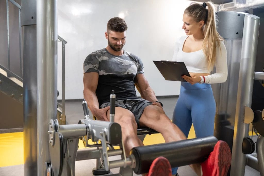 A trainer takes notes as a client performs reps on a leg extension machine.