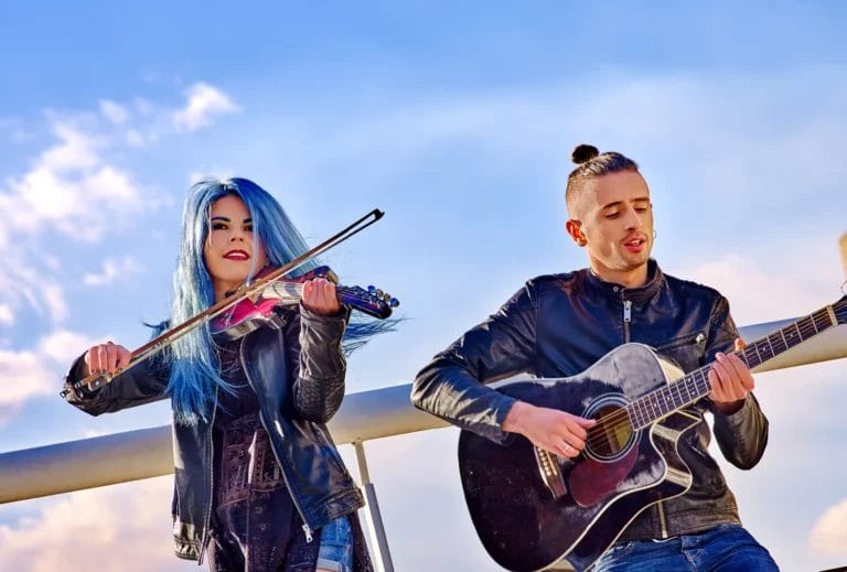 Two musicians performing outdoors, one playing violin and the other playing guitar, with a bright sky in the background.