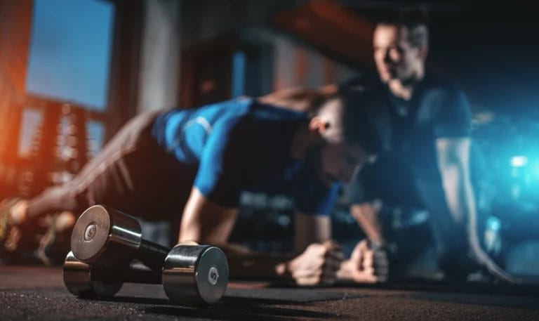 A dark gym, with a set of dumbbells in the foreground and an out-of-focus trainer working with a client in the background.