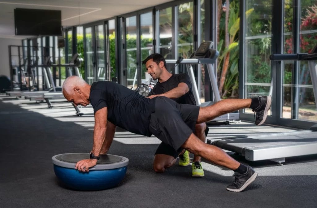 A personal trainer guides an older client through a plank exercise with a Bosu ball.