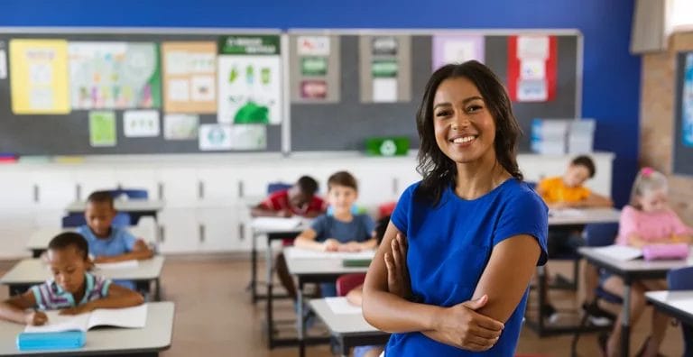 A teacher in a blue shirt and white pants leans against a desk and smiles at the camera in front of her classroom full of elementary school students.