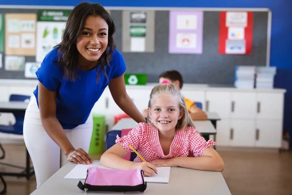 A teacher wearing a blue shirt and white pants stands next to her student seated at a desk and wearing a pink shirt. Both teacher and student are smiling at the camera.