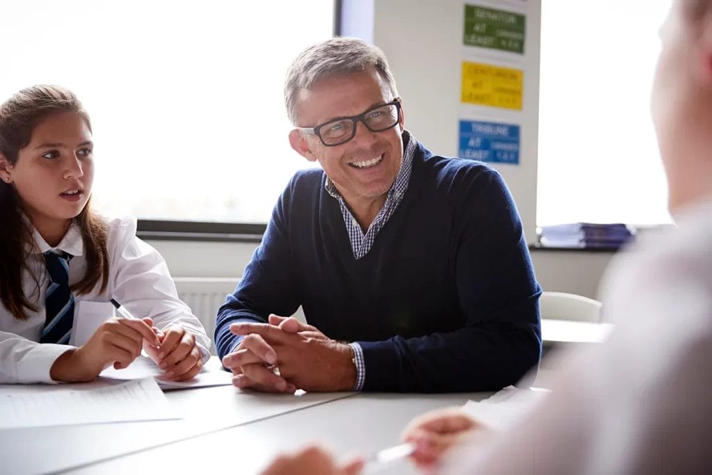 An older male teacher wearing glasses and a blue sweater sits at a desk in a well-lit classroom with two private school students, smiling at one of them.