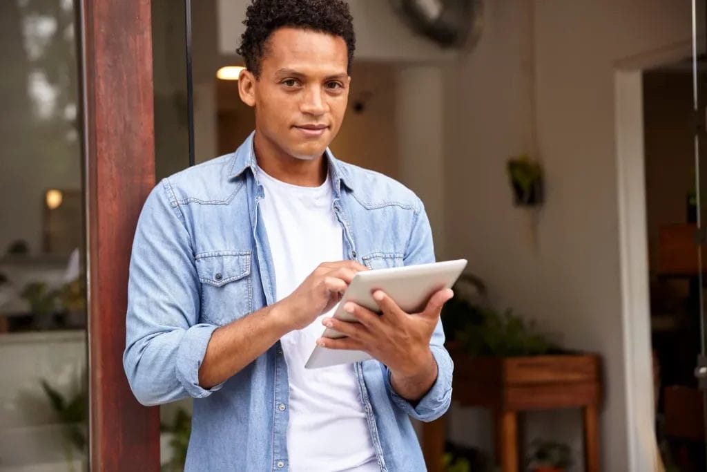 A small business owner wearing a denim shirt smiles and taps on his business tablet while leaning against a doorframe with white walls and green plants in the background.