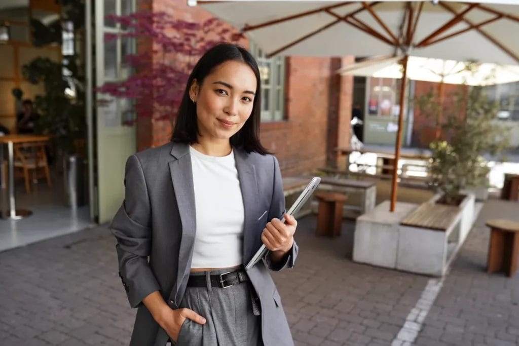 A small business owner wearing a gray suit and white shirt holds a business tablet and smiles outside a red-brick building with a white canopy outdoor seating area.
