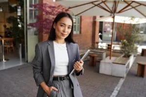 A small business owner wearing a gray suit and white shirt holds a business tablet and smiles outside a red-brick building with a white canopy outdoor seating area.
