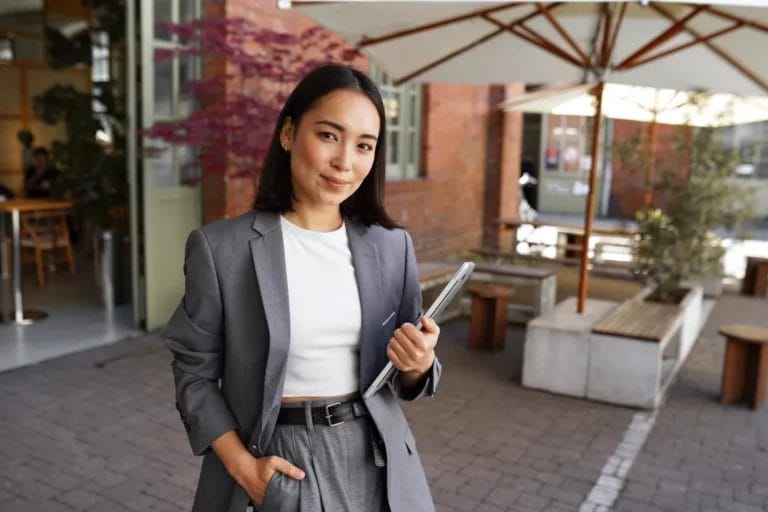 A small business owner wearing a gray suit and white shirt holds a business tablet and smiles outside a red-brick building with a white canopy outdoor seating area.