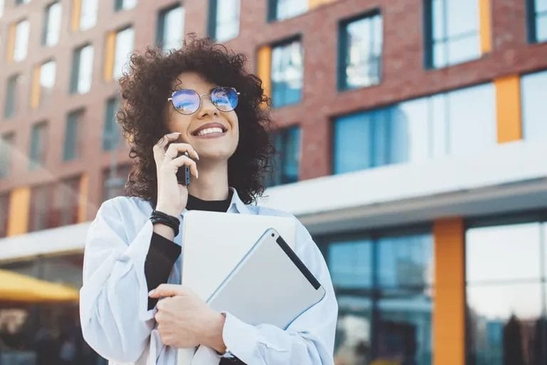 Smiling woman wearing glasses and holding a laptop while talking on a cellphone