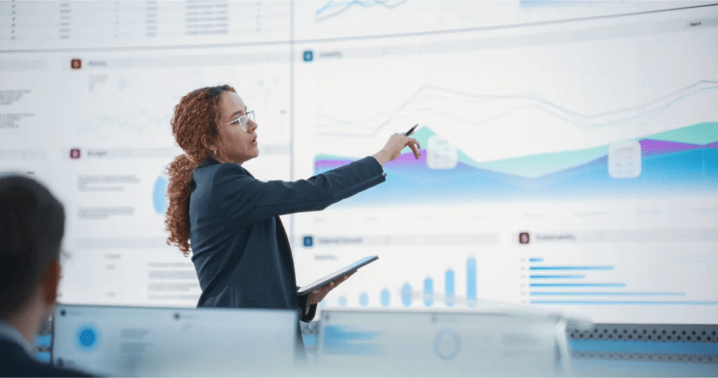 Consultant points to a graph and data on a large screen behind her
