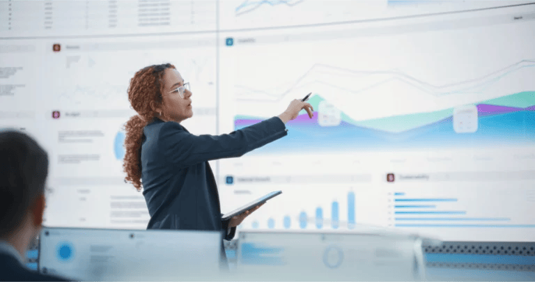 Consultant points to a graph and data on a large screen behind her