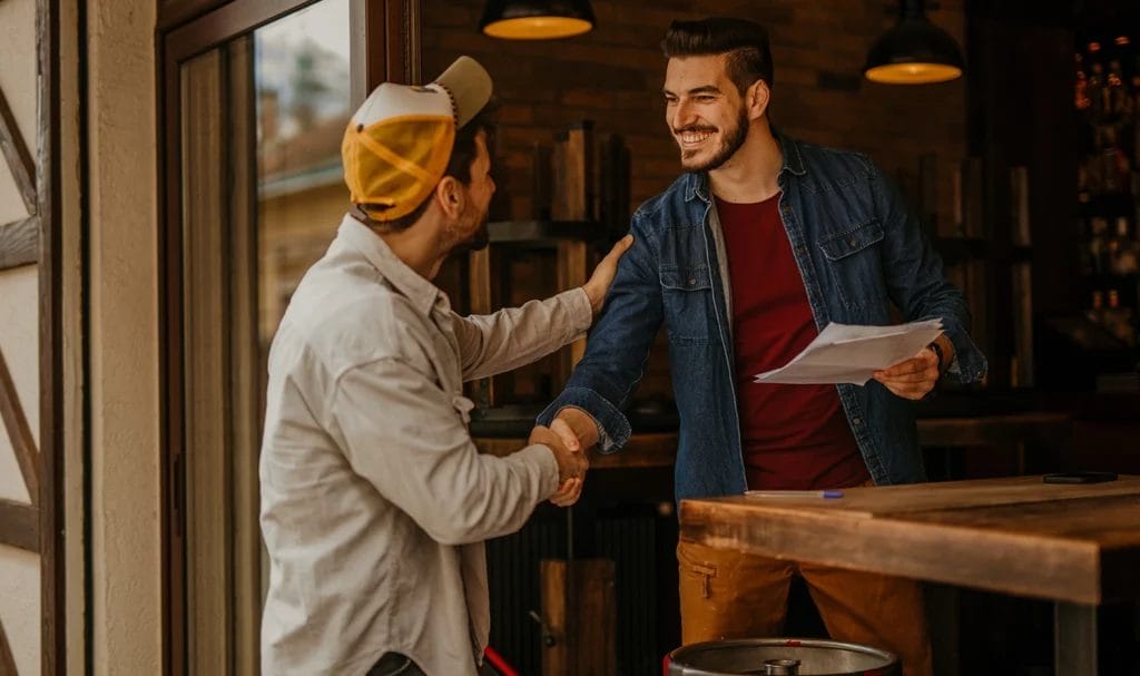 Two business owners happily shake hands as one holds a few documents in his other hand.