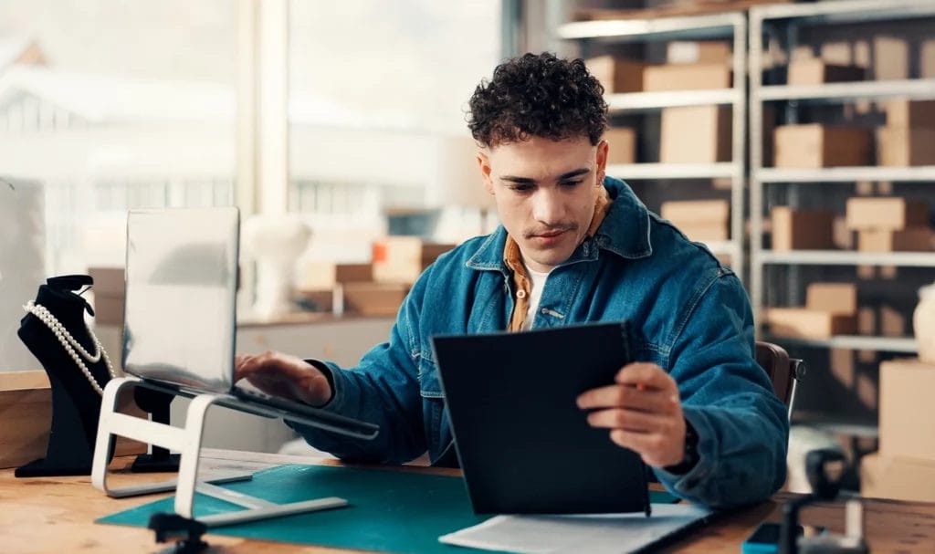 A young entrepreneur uses a tablet and a laptop in a small office space surrounded by shelves of ready-to-ship product boxes.