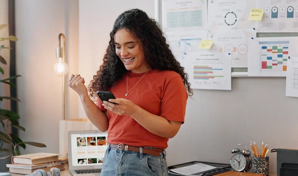 A small business owner celebrates in her home office as she reads the news of a new deal secured for her brand on her smartphone.