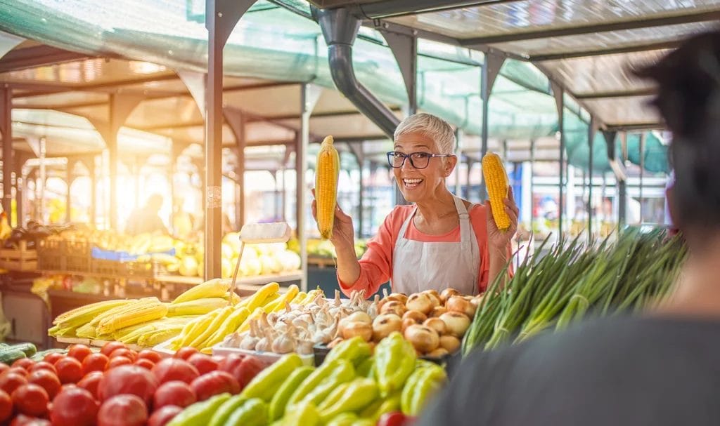 A farmers market vendor happily smiles at the camera from her both as she excitedly waves to ears of corn in the air.
