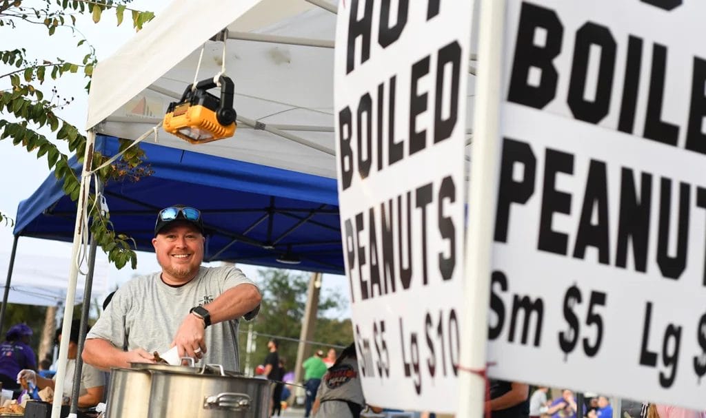 A peanut vendor happily smiles for the camera as he preps a pot of boiled peanuts at an outdoor event.