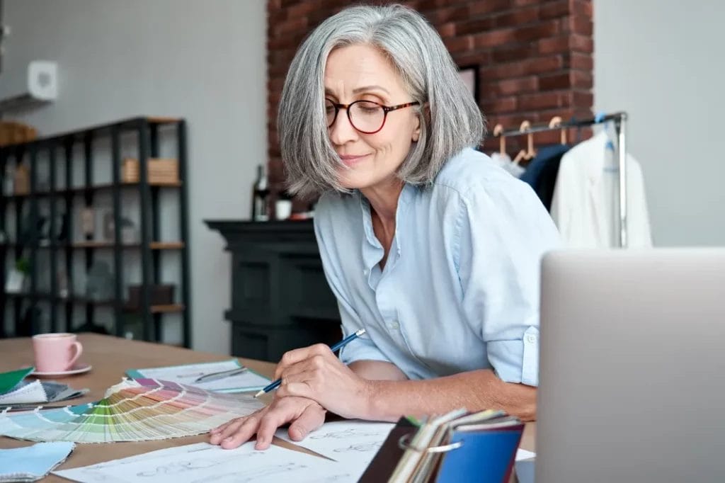 A small business owner wearing a light blue button-up shirt looks over documents while sitting at a wooden desk in a design studio.