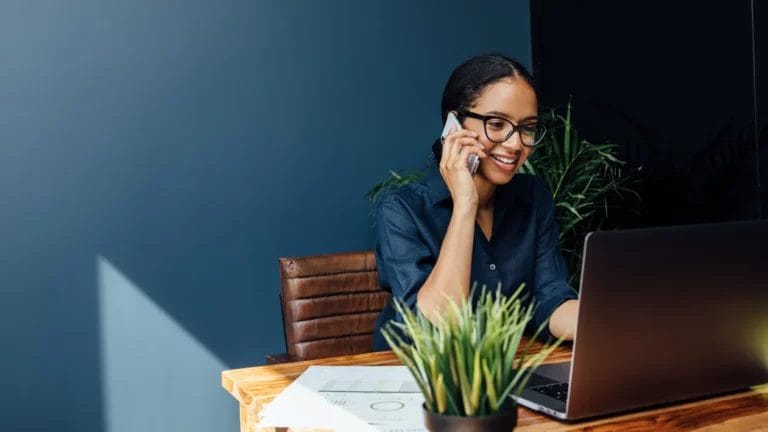 A small business owner wearing a teal button-up shirt sits at a wooden desk and speaks on her smartphone while looking at a gray laptop in a teal-colored room.