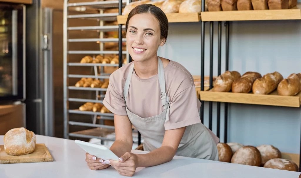 A baker smiles at the camera while holding a tablet.