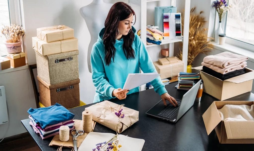 A young ecommerce business owner is managing orders from her laptop in her home office, surrounded by packages, samples, products, and shipping supplies.