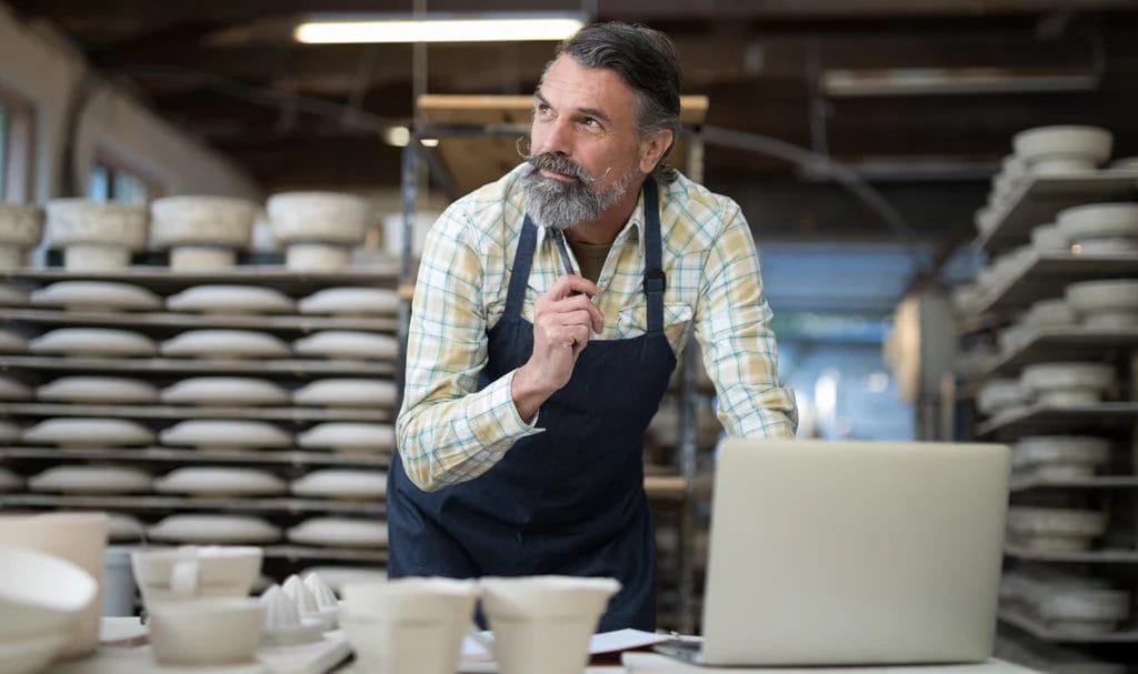 A mustachioed potter smiles as he looks in the distance in thought while using a laptop in his pottery studio.