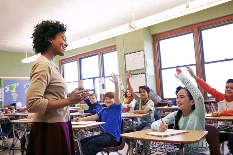 A female teacher stands in front of a classroom of late elementary school students with their hands raised.