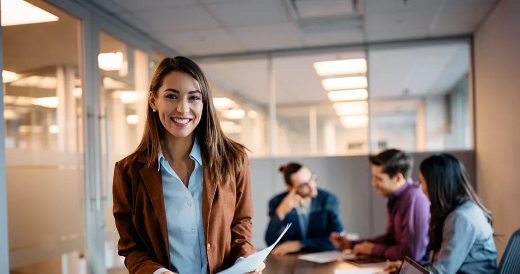 A young woman smiles at the camera wearing a blue button-up shirt and brown blazer in a corporate conference room with three coworkers in the background.