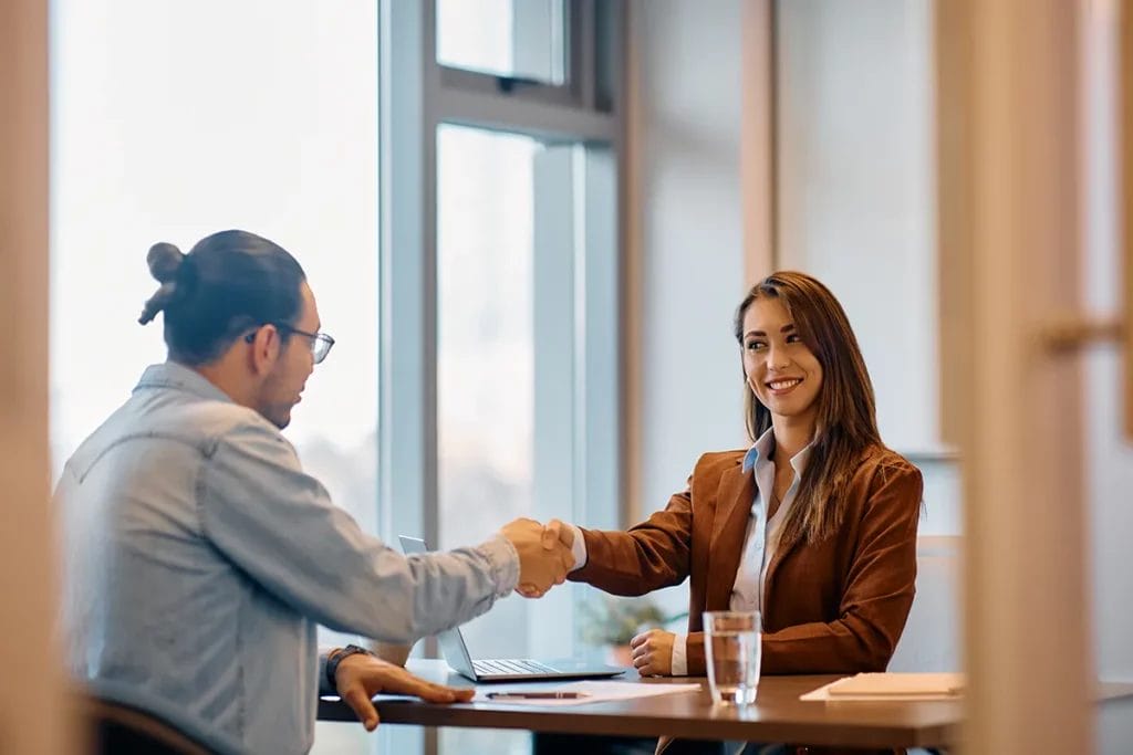 A woman in a brown blazer shakes hands across a table with a man in a denim shirt in a corporate conference room.