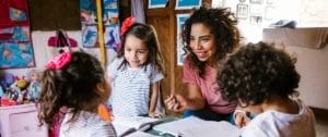 A woman sits at a small table with three children who have workbooks open in front of them.