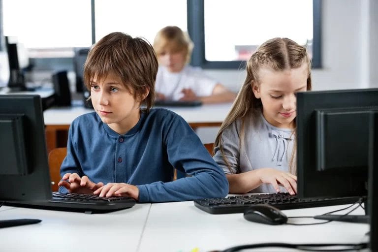 Two students, a boy and a girl, use desktop computers during class in a brightly lit computer lab.