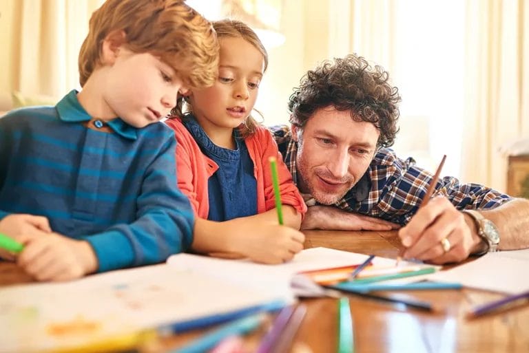 A man with curly hair draws something with a colored pencil alongside two young children in a homeschool environment.
