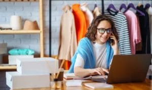 Small business woman chatting on the phone while working on her laptop with custom clothing hanging behind her.