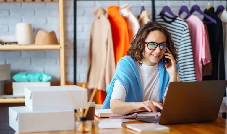 Small business woman chatting on the phone while working on her laptop with custom clothing hanging behind her.