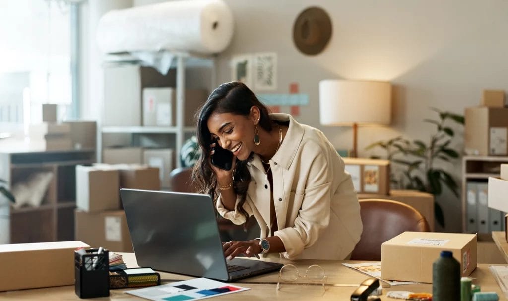A businesswoman is smiling while talking on her phone and typing on her laptop as she leans over her desk in her home office, surrounded by packages and shipping supplies.