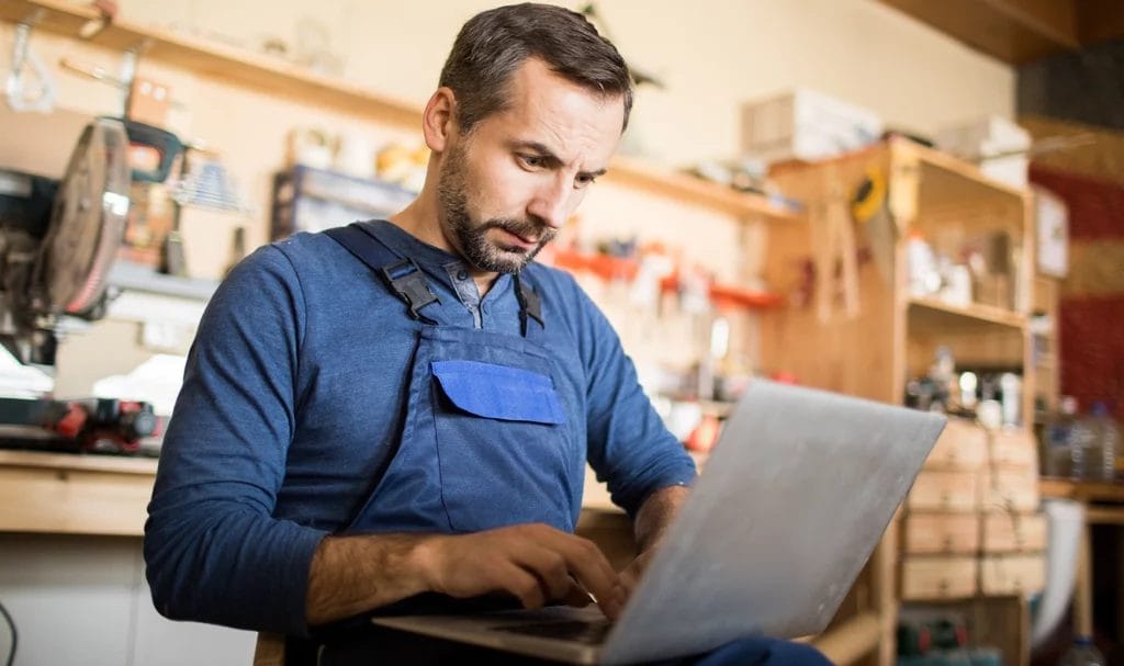 A woodworker sits in his shop, working on his laptop.