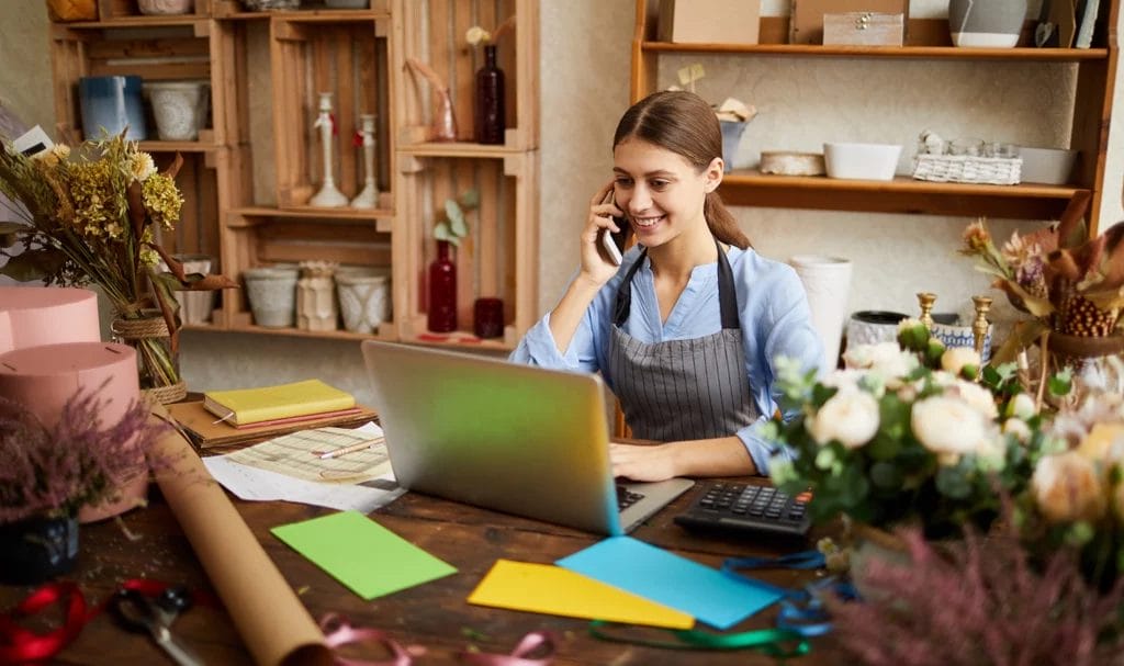 A small business florist smiles as she speaks on the phone and looks at her laptop, sitting at a workbench in her studio.