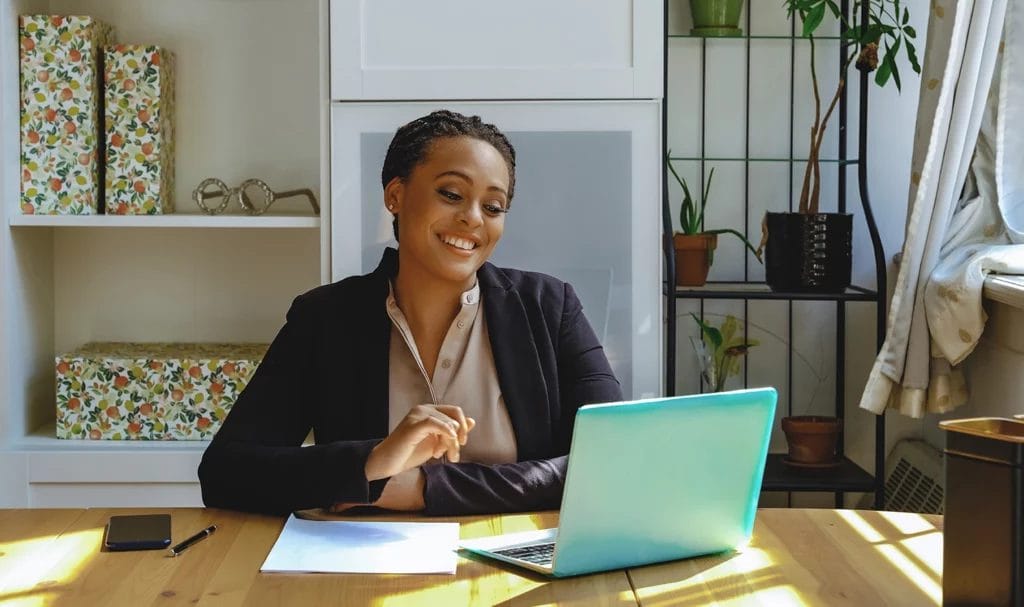 A small business owner happily works from a home office on a teal laptop as she smiles at her computer screen.