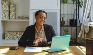A small business owner happily works from a home office on a teal laptop as she smiles at her computer screen.
