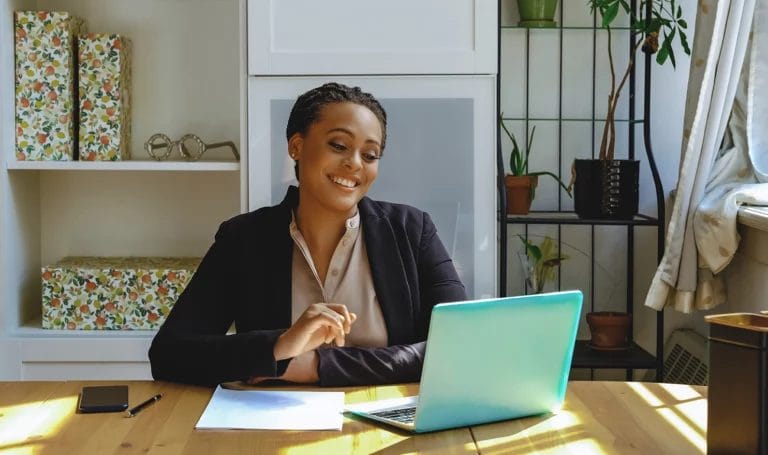 A small business owner happily works from a home office on a teal laptop as she smiles at her computer screen.